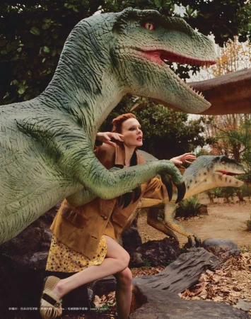Woman poses dynamically with dinosaur statues outdoors, showcasing a floral dress and jacket in a staged editorial environment.
