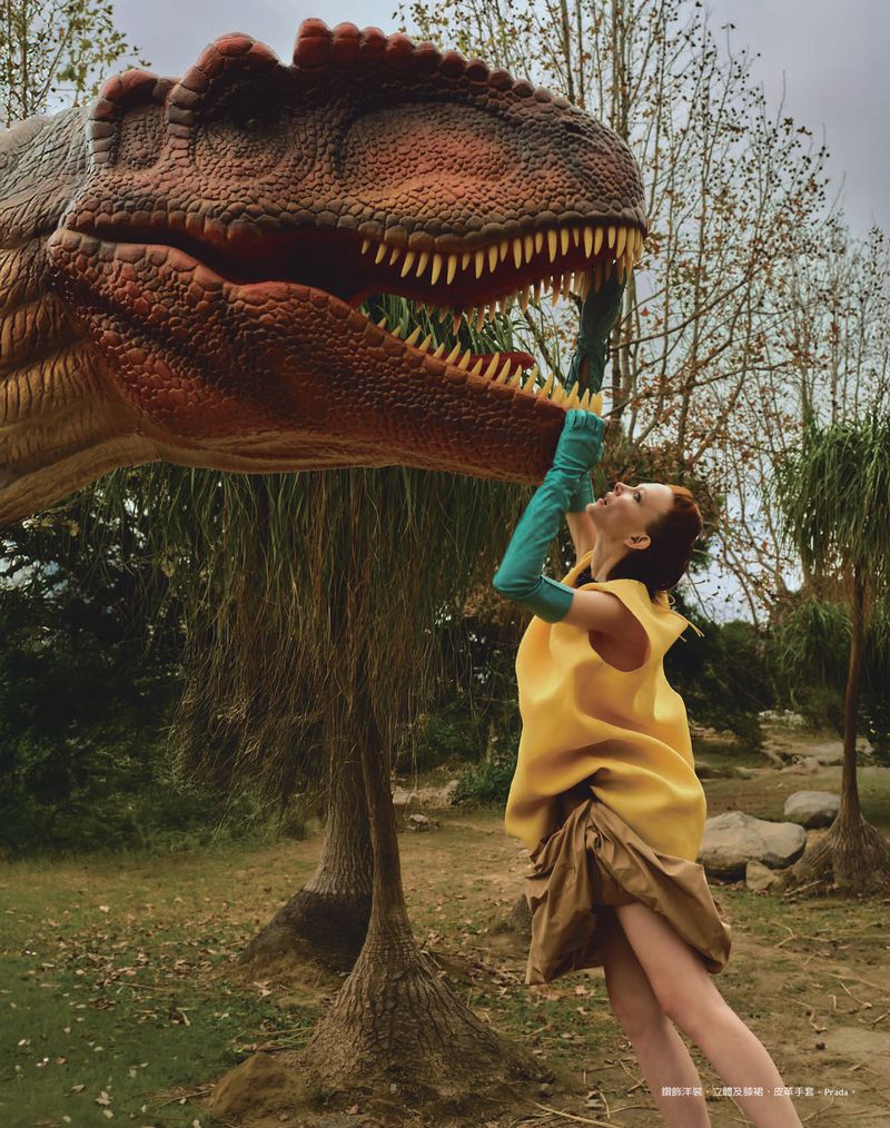 Woman in yellow dress and green gloves reaches to touch a dinosaur head in an outdoor park-like setting, trees and lawn visible.