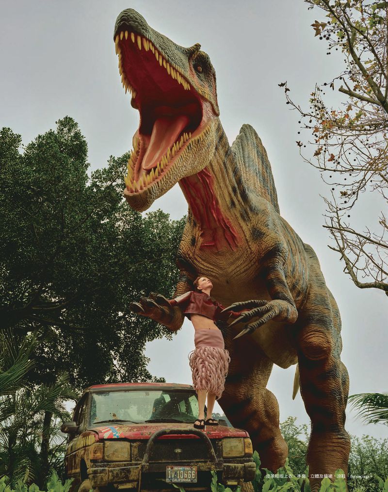 A woman stands on a damaged jeep in front of a dinosaur statue in an outdoor setting with trees and foliage, capturing a unique scene.