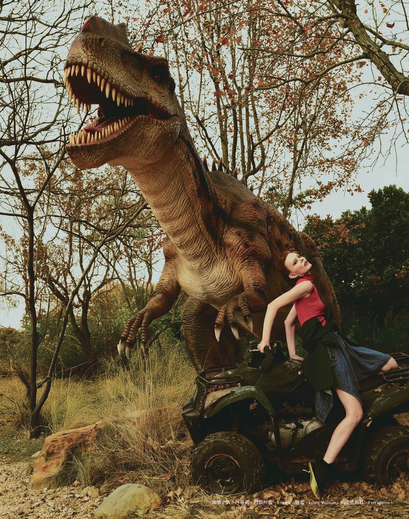 A fashion image featuring a woman posing on an all-terrain vehicle with a large dinosaur model in a wooded, outdoor environment.