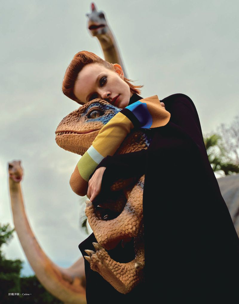 Adult woman hugs a toy dinosaur against a sky background with brachiosaurus statues in a fashion editorial style medium shot.