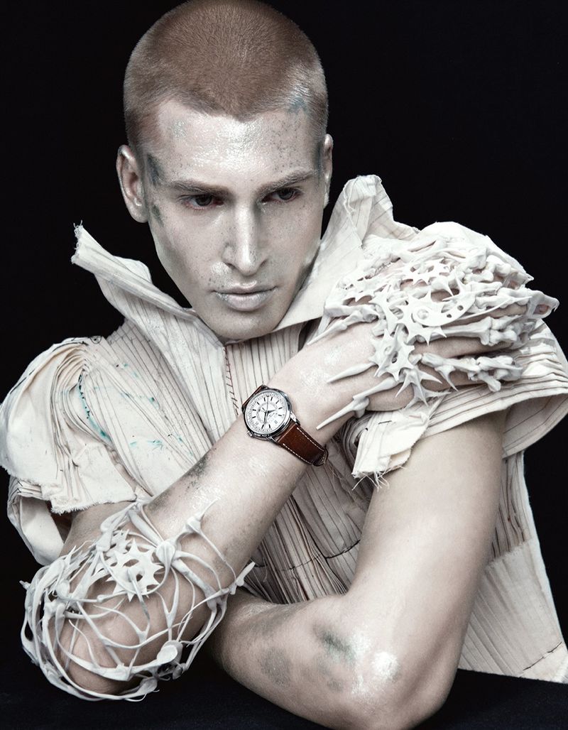 Studio portrait of a man with painted pale skin and a shaved head, wearing a ruffled collar and sculptural sleeves, and a wristwatch.