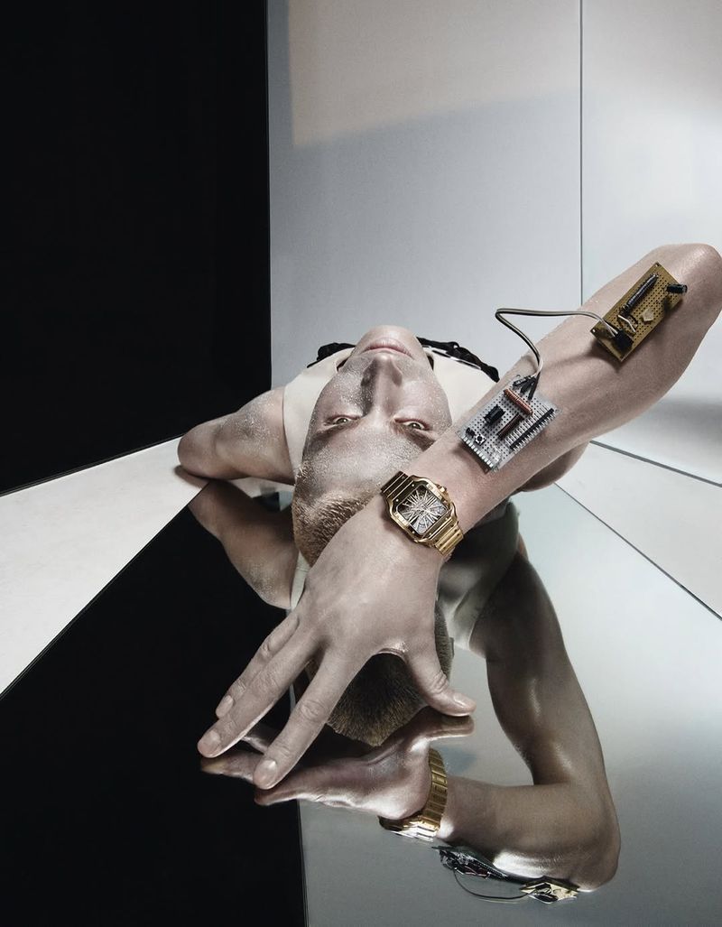 Upside-down view of adult male model in studio with gold watch, silver and gold electronics, and black-and-white background.