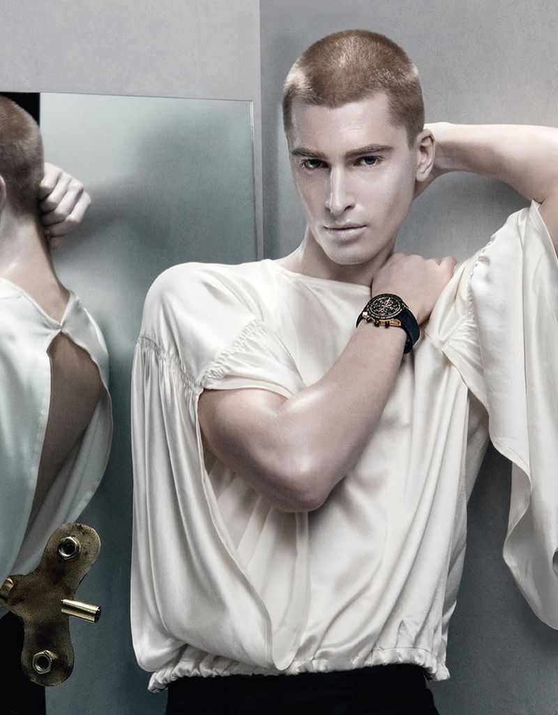 Androgynous person with a shaved head poses in front of a mirror, wearing a white blouse and a watch, with a toy wind-up key.