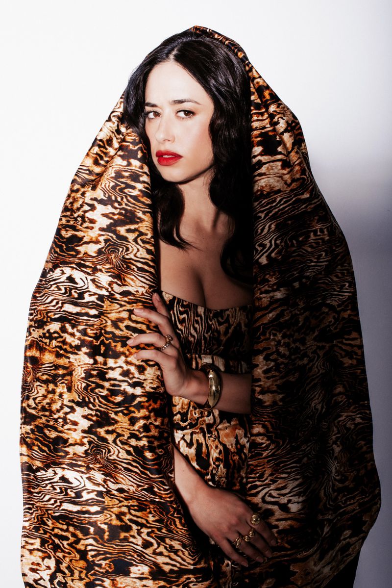 Portrait of Jeanine Mason wearing a patterned hooded garment and strapless dress against a white studio background.