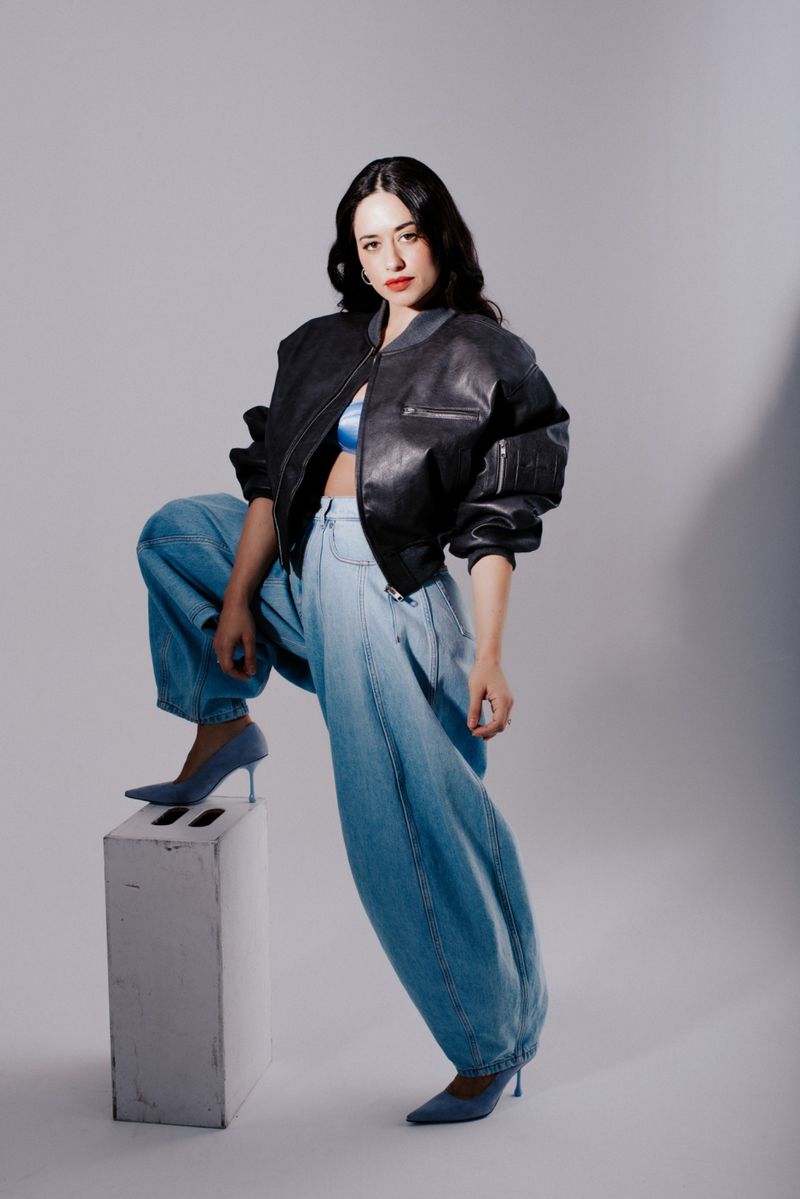 Adult woman poses in a studio wearing a black leather bomber jacket, blue top, baggy jeans, and blue heels while standing on a block.