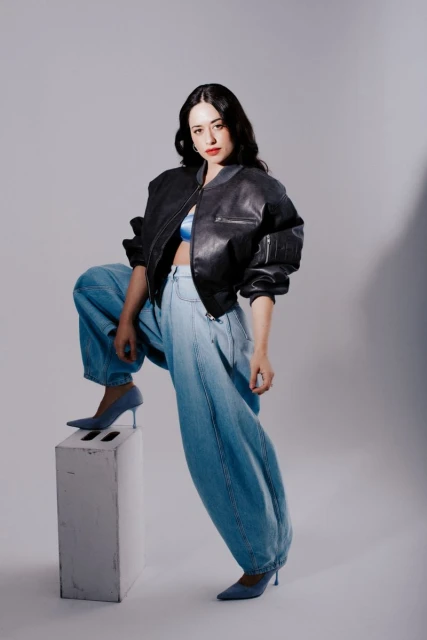 Adult woman poses in a studio wearing a black leather bomber jacket, blue top, baggy jeans, and blue heels while standing on a block.