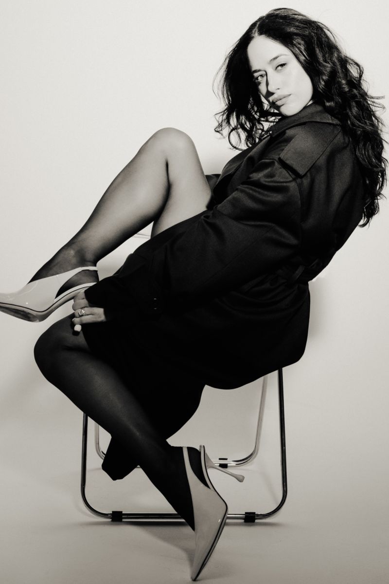 Black and white shot of a woman in a dark coat, pantyhose, and heels, sitting on a chair in a photo studio setting, looking at the camera.