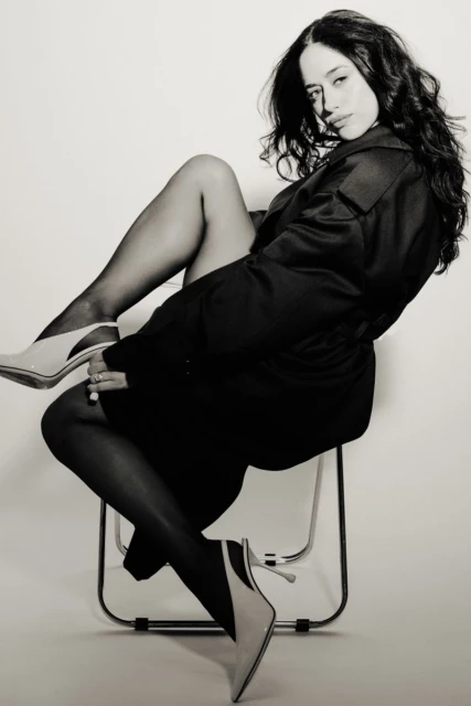 Black and white shot of a woman in a dark coat, pantyhose, and heels, sitting on a chair in a photo studio setting, looking at the camera.