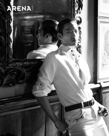 Black and white portrait of an adult male model leaning against a decorative wall with a mirror, wearing a white shirt and black belt.