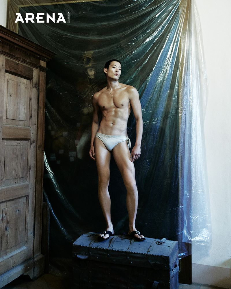 Asian man stands on a trunk in swimwear in an indoor setting with an old cabinet on the left and draped plastic sheeting in the background.