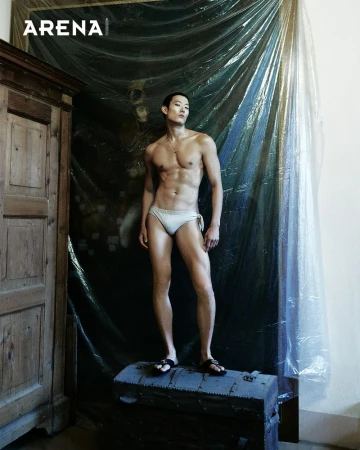 Asian man stands on a trunk in swimwear in an indoor setting with an old cabinet on the left and draped plastic sheeting in the background.