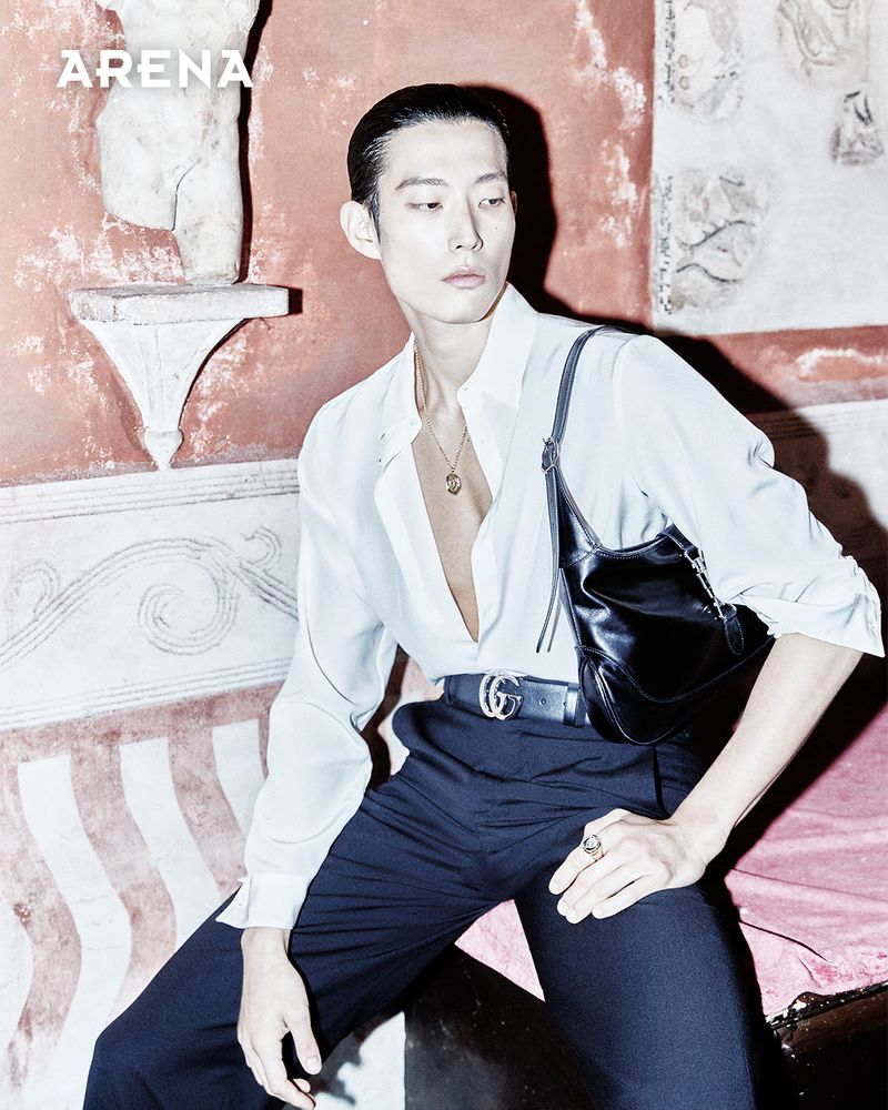 Asian male model poses in a white shirt and black pants with a Gucci belt and black bag in front of an ancient wall for fashion.