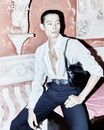 Asian male model poses in a white shirt and black pants with a Gucci belt and black bag in front of an ancient wall for fashion.