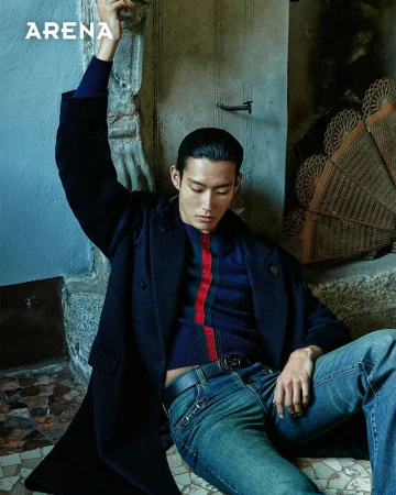 An Asian male model is sitting on stone steps in a dark blue coat and blue jeans, holding his arm up in a fashion editorial shoot.
