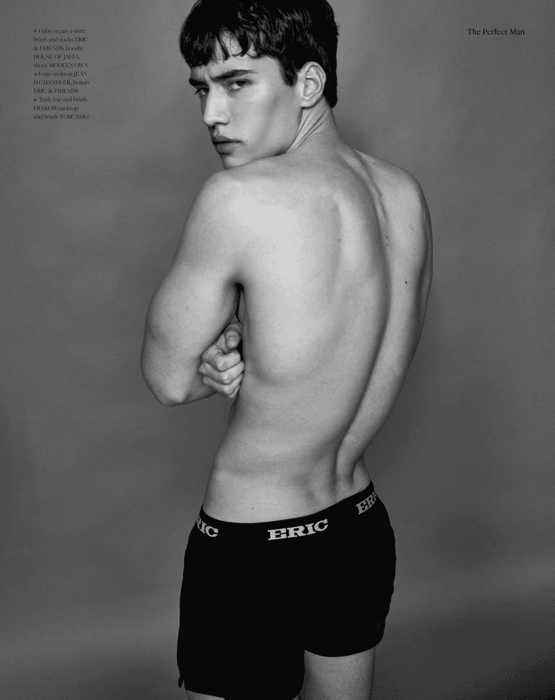 A shirtless young man with dark hair poses against a gray background, wearing black boxer shorts. The image is black and white.
