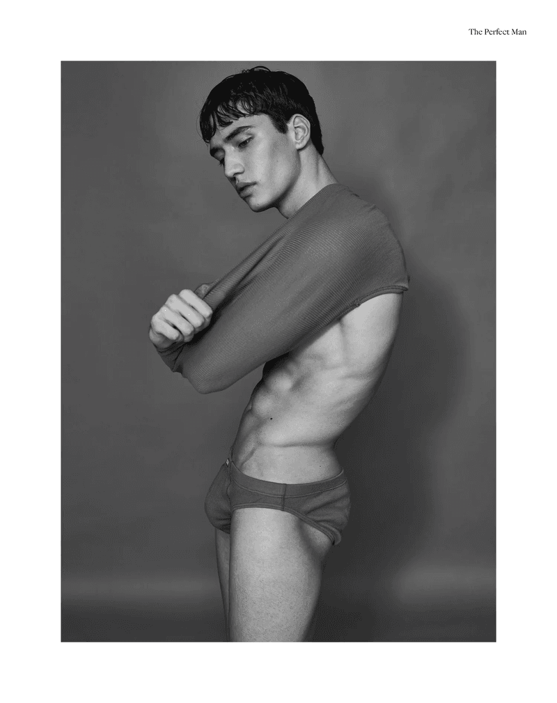 Young man with short dark hair in profile, pulling up a fitted rib-knit top, wearing briefs against a plain grey studio background.