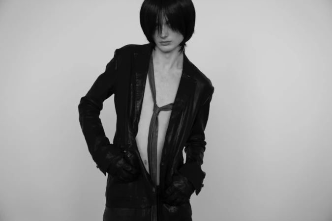 A person with dark hair poses wearing a dark jacket and tie against a plain white background in a studio shot with a fashion style.