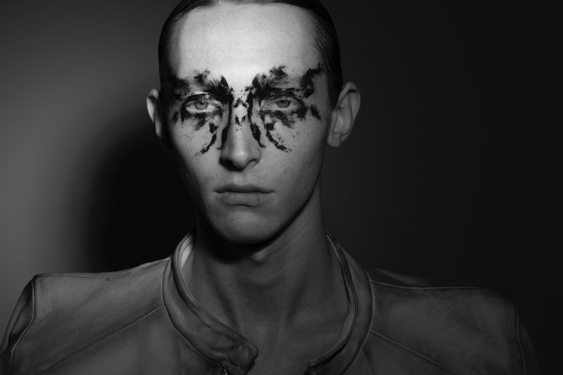 Close-up portrait of a young adult with decorative black face paint, set against a neutral gray backdrop in a monochromatic composition.