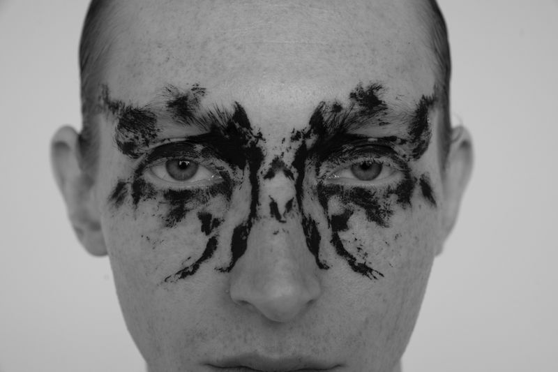 Close-up black-and-white studio portrait of a person's face with dramatic graphic black makeup around the eyes in an abstract pattern.