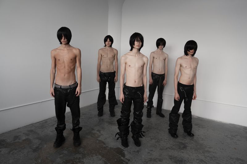 Group of five slender, shirtless young men with dark wigs and black pants pose in a sparse studio with white walls and a gray floor.