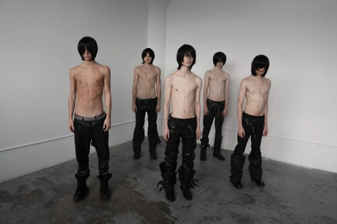 Group of five slender, shirtless young men with dark wigs and black pants pose in a sparse studio with white walls and a gray floor.