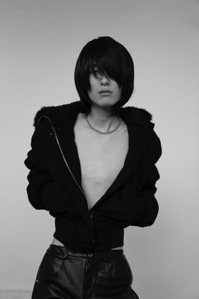 Monochrome studio portrait of a person with a dark bob haircut wearing an open black zip-up jacket and shiny black trousers.
