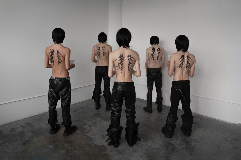 Five shirtless young adults with drawings on their backs wear black pants in a minimalist white interior, showcasing fashion and body art.