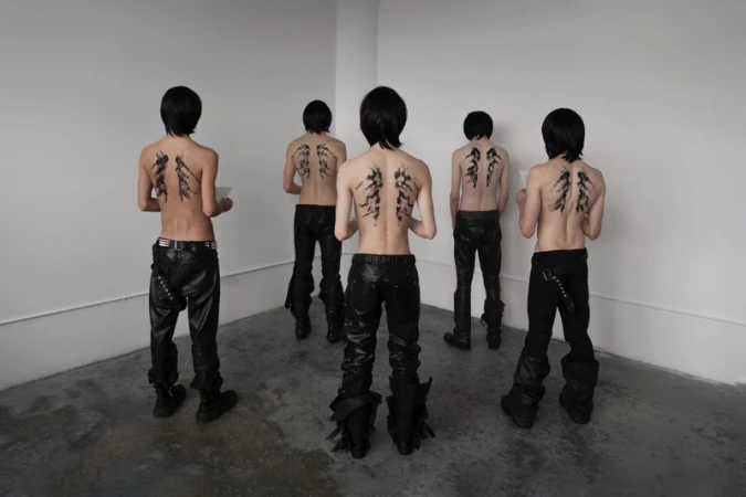 Five shirtless young adults with drawings on their backs wear black pants in a minimalist white interior, showcasing fashion and body art.