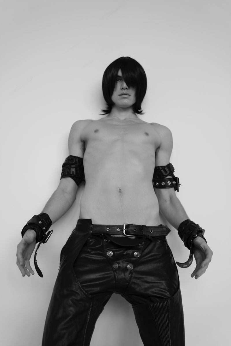 Shirtless man with dark hair poses in black leather pants and studded wristbands against a white wall in black and white.