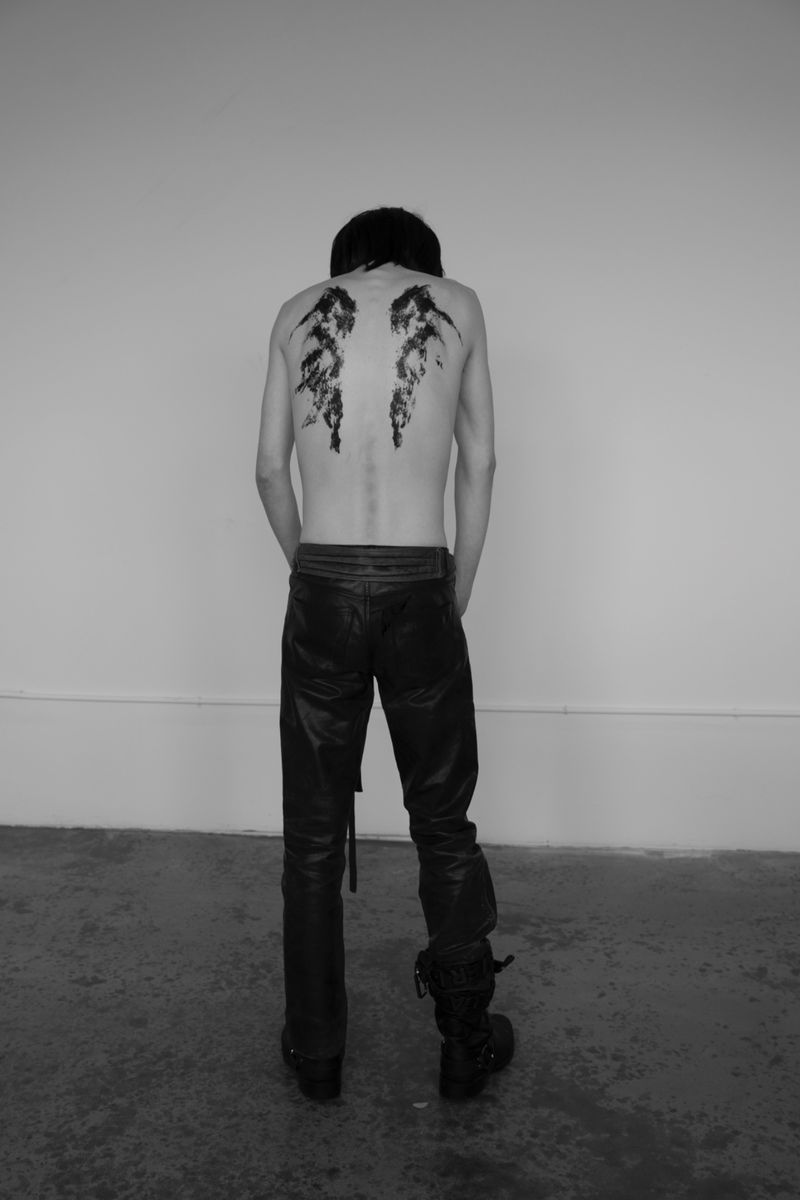 Monochrome back view of person with painted wings on their back, wearing black leather pants and boots, standing against a white wall.