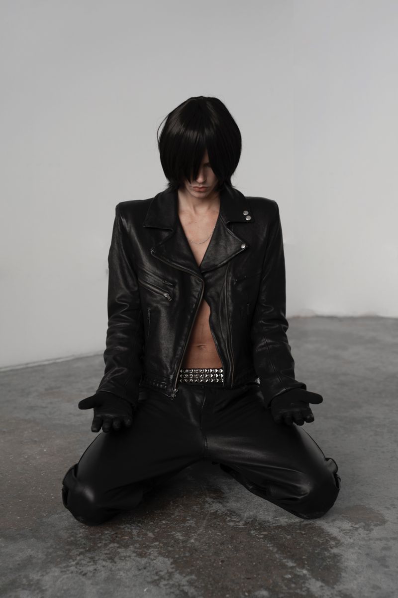 Person kneels in black leather jacket and pants, head bowed, in a stark white studio. Minimalist fashion photography focusing on texture and form.