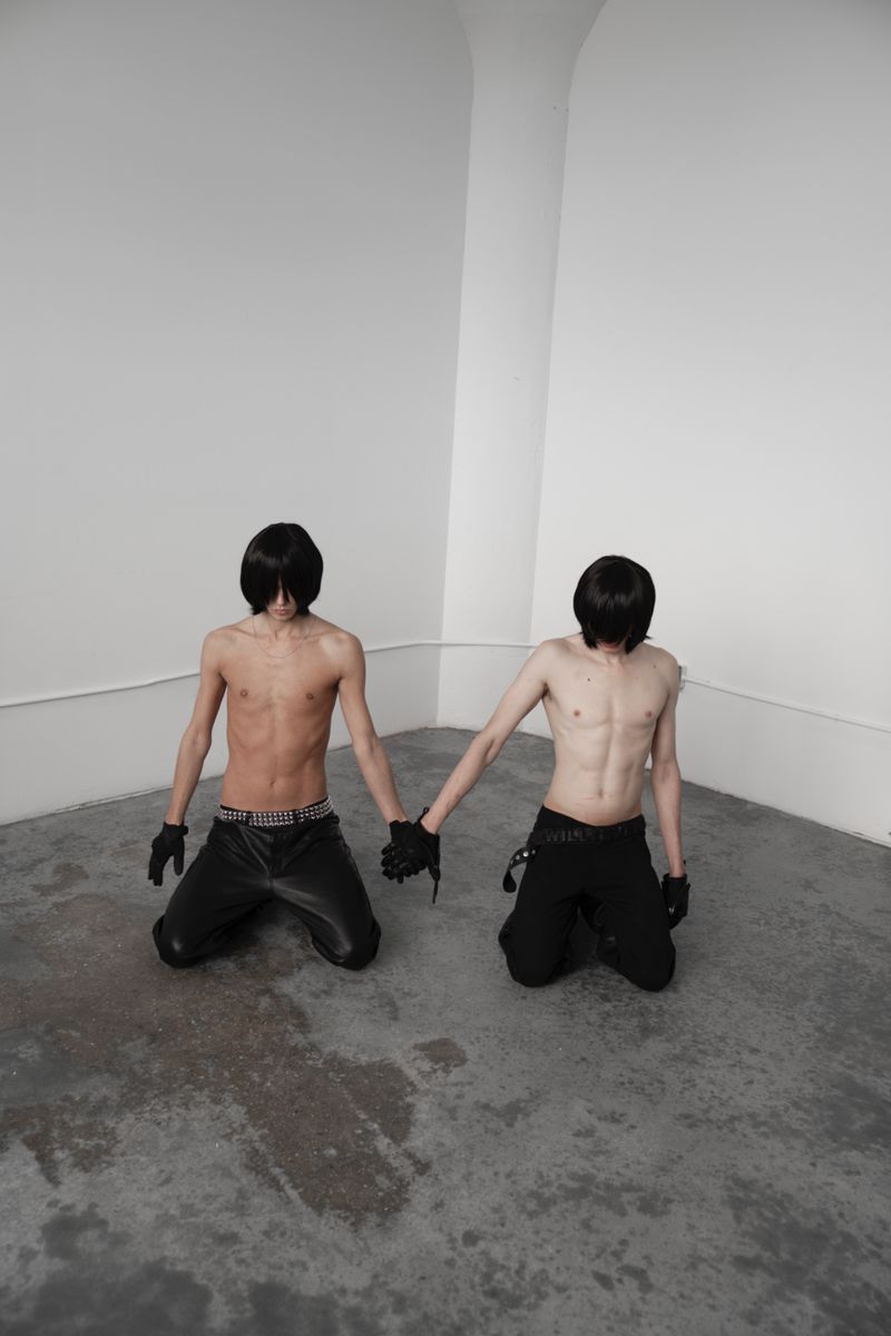 Two shirtless, kneeling men in black pants and gloves hold hands in a white corner, creating an evocative fashion or fine art image.