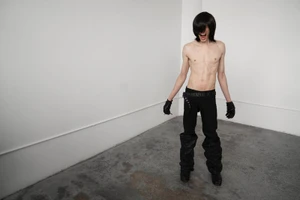 Shirtless man with dark hair over his face wearing black trousers and leather gloves in an empty white room