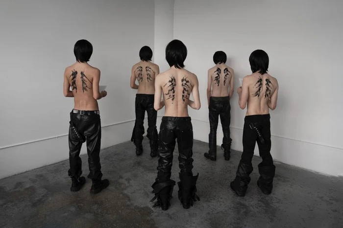 Five shirtless models standing with their backs to the camera wearing black leather trousers in a minimal room.