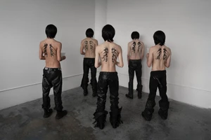 Five shirtless models standing with their backs to the camera wearing black leather trousers in a minimal room.