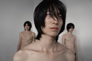 Close-up portrait of a dark-haired male model with two shirtless models standing in the blurred background against a white wall.