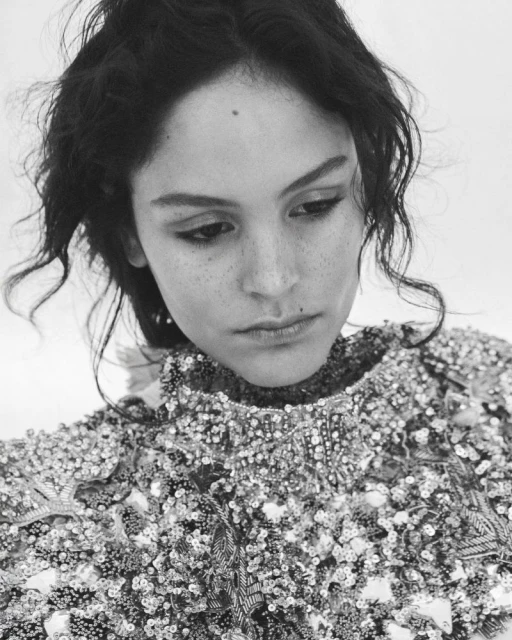 Monochrome portrait of a young woman with dark curly hair wearing a sequined top, looking down with a subdued expression.