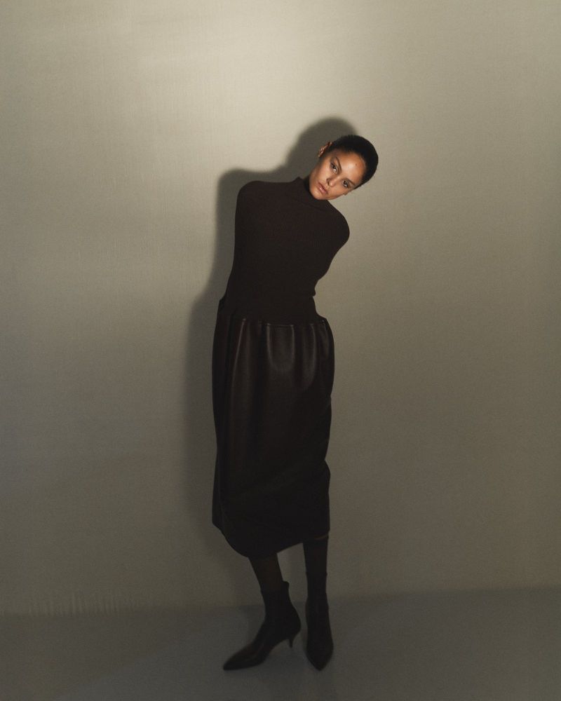 A person with short hair wearing a long-sleeved dark brown turtleneck and a dark skirt poses against a pale grey wall with hard shadows.