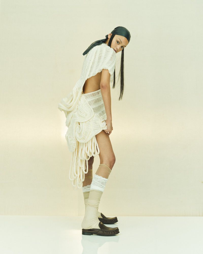 Fashion photo of a young adult woman in a cream dress with rope details, leather head covering, boots, in a studio setting.