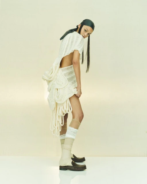 Fashion photo of a young adult woman in a cream dress with rope details, leather head covering, boots, in a studio setting.