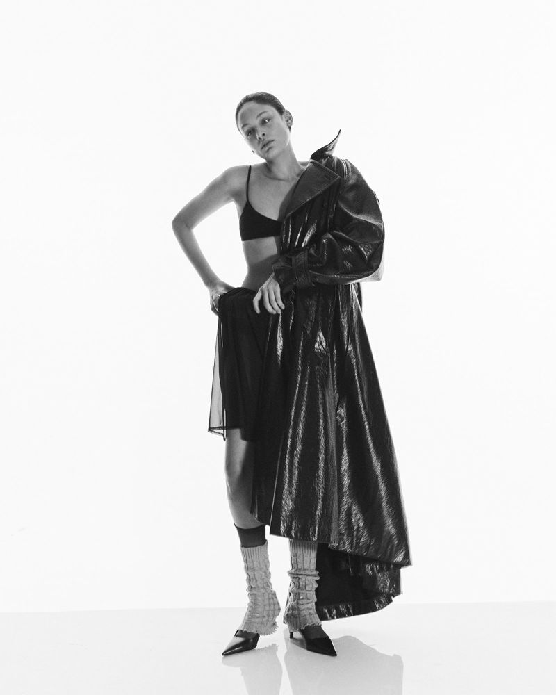 Fashion shot in black and white of a slender woman wearing a black bra, dress, coat and leg warmers in a bright, seamless studio.