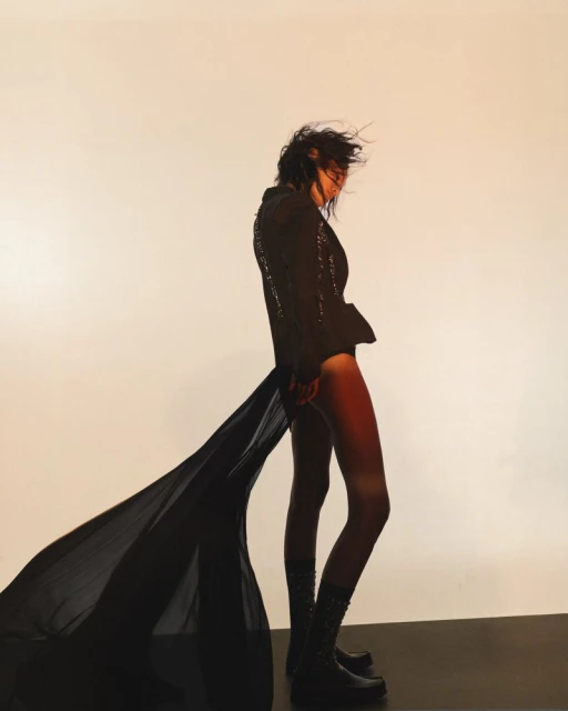 Woman posing in a black blazer with a long, flowing train and ankle boots against a neutral gradient background, fashion shot.
