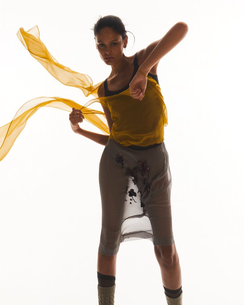A model poses with a translucent yellow scarf, wearing a yellow top and grey embellished skirt against a bright white background in studio.