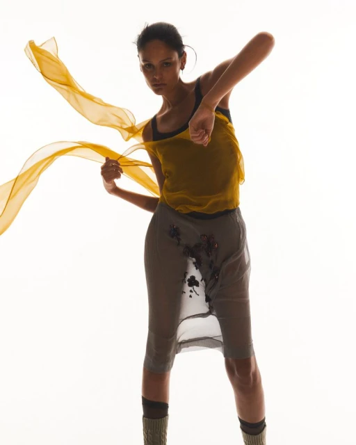 A model poses with a translucent yellow scarf, wearing a yellow top and grey embellished skirt against a bright white background in studio.