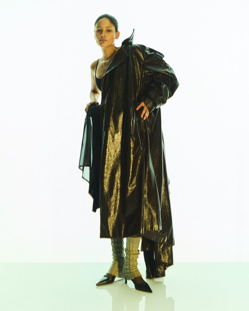 Fashion editorial shot of a Black woman posing in a black bra top and a shimmering trench coat against a bright white background.