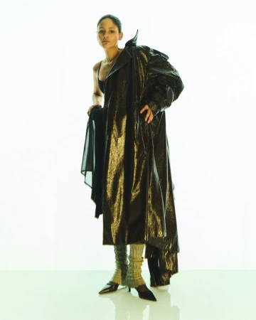 Fashion editorial shot of a Black woman posing in a black bra top and a shimmering trench coat against a bright white background.