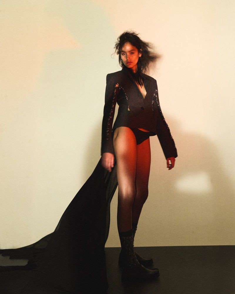 A woman poses in a black blazer and underwear against a gradient background, casting a shadow on the floor in a fashion style photo.