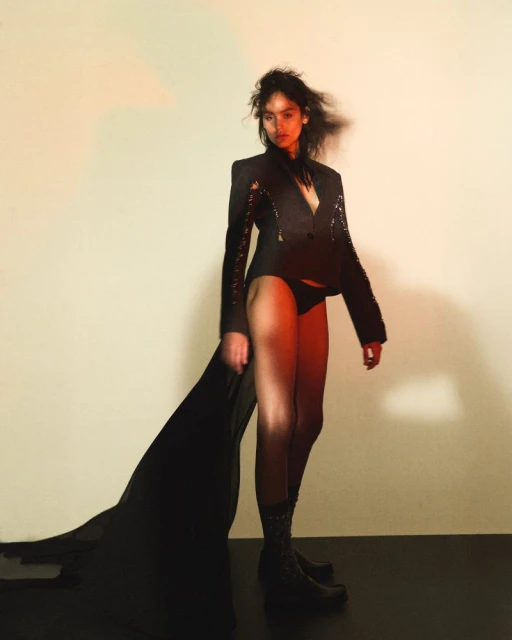 A woman poses in a black blazer and underwear against a gradient background, casting a shadow on the floor in a fashion style photo.