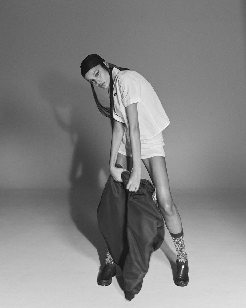 Young woman in a white T-shirt and shorts poses in a studio with a black jacket, patterned socks, and black shoes against a grey backdrop.